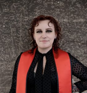 Woman wears a red graduation sash over a black blouse. She has grey eyeshadow