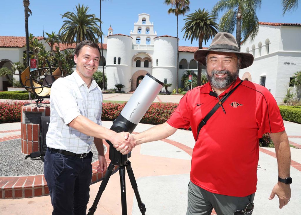 Tom Esposito (left) shakes hands with Bill Welsh (right) with the Unistellar eVscope 2 and Hepner Hall in the background