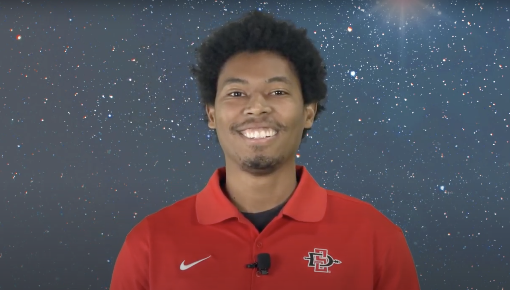 A young man wearing a red SDSU polo shirt smiles in front of a space virtual background