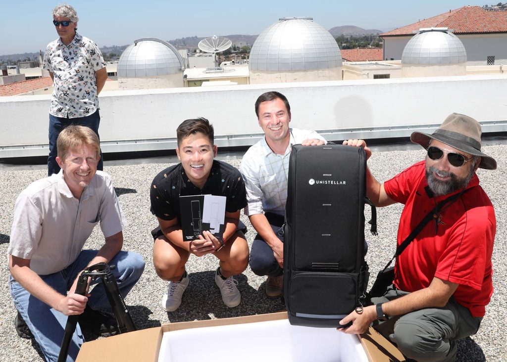 Eric Sandquist, Thomas Tran, Thomas Esposito and Bill Welsh unbox the Unistellar telescope with the observatory domes and Associate Dean Tod Reeder in the background.