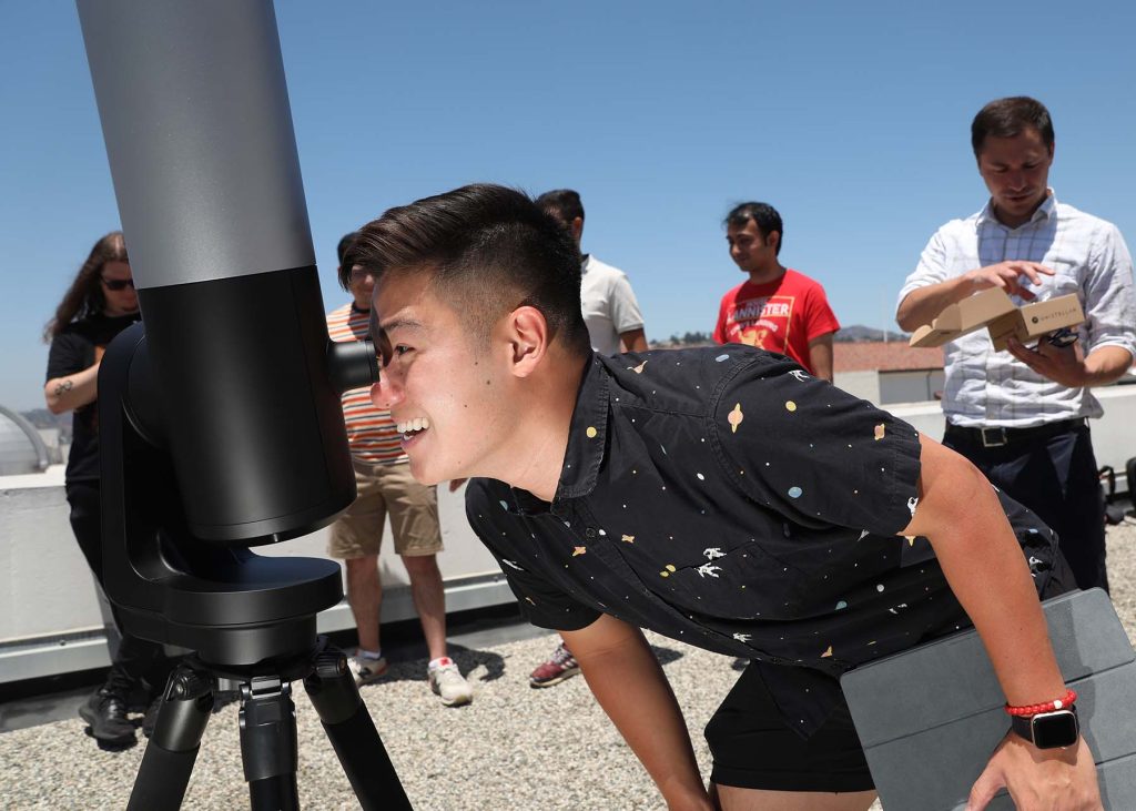 Thomas Tran wearing an astronomy themed black shirt looks through the eyepiece of the telescope at the sun