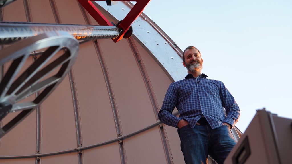 A man in a blue plaid button-up shirt with a beard stands inside of a telescope observatory dome at sunset