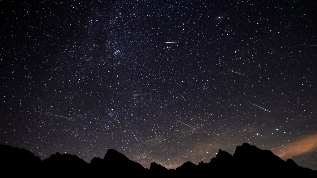 Dozens of long white streaks trail after stars against a dark background littered with bluish white stars above the silhouette of mountains. Photo from Adobe Stock