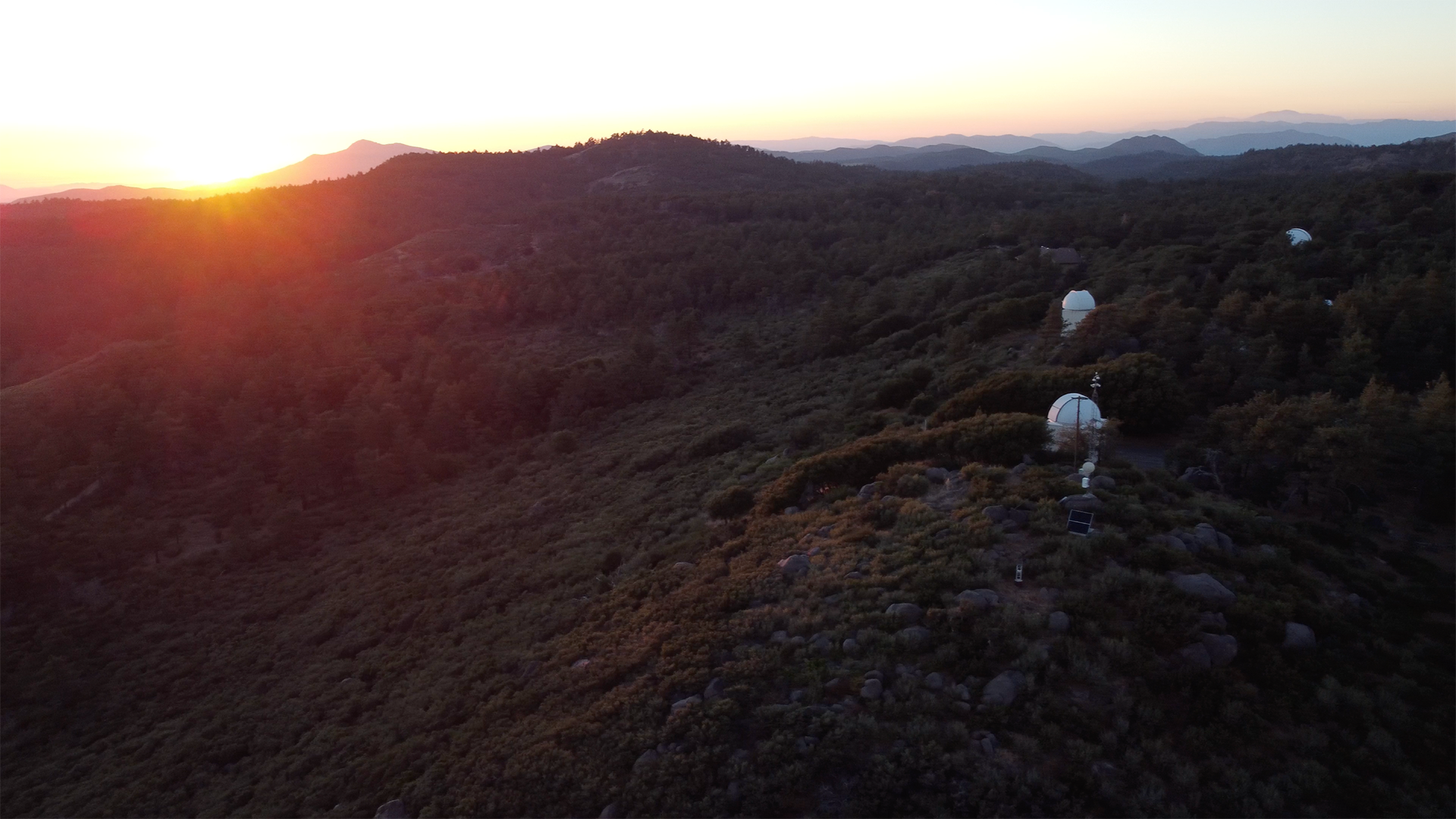 Wooded mountains at sunset with three white observatory towers visible amongst the trees